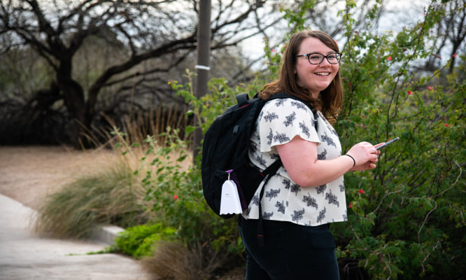 adult walking outdoors holding a phone and wearing a backpack with the air sensor attached to it.